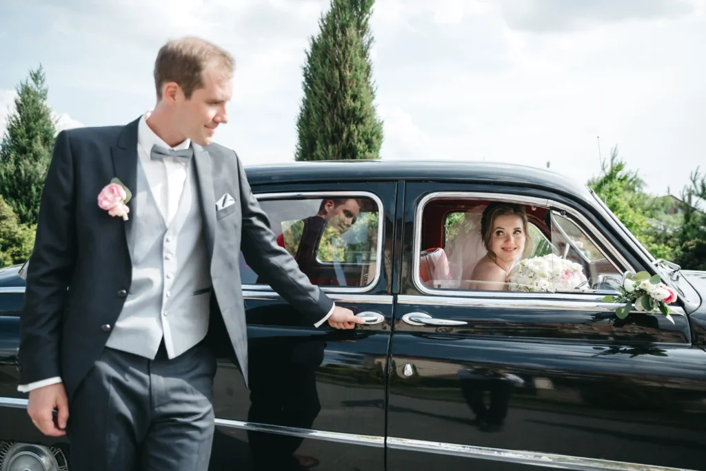 Professional event chauffeur service with groom opening a luxury vintage car door for bride on wedding day